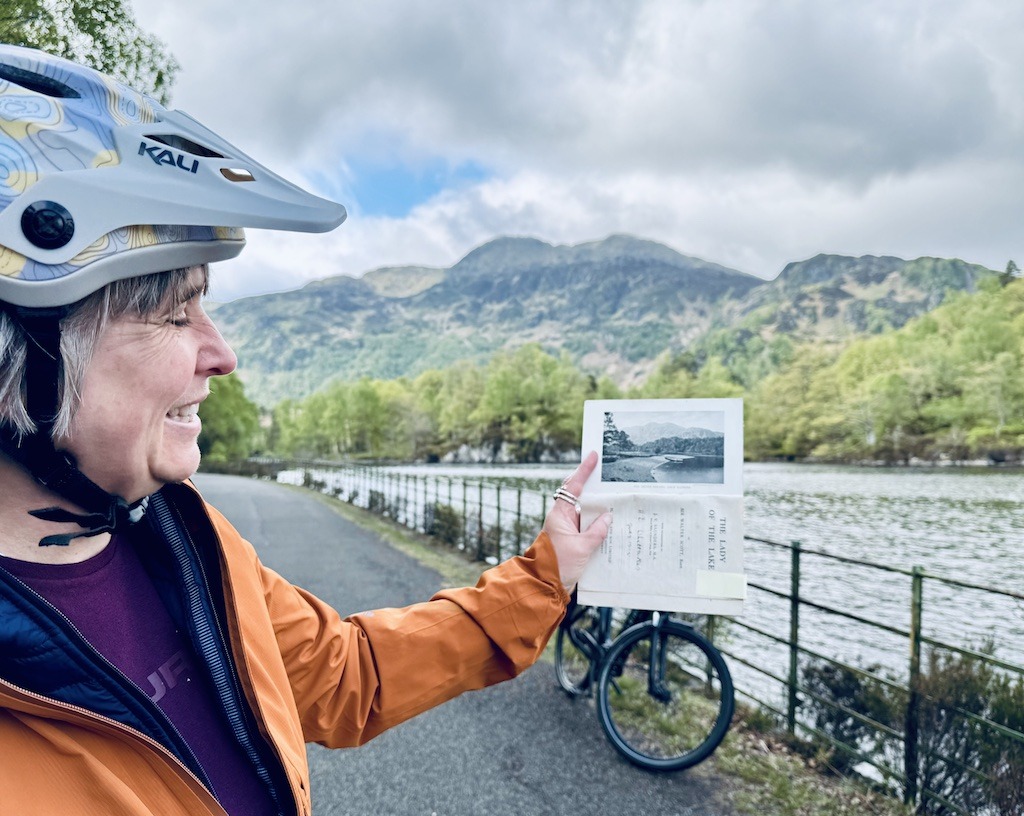 woman in orange jacket and bike helmet holding up a book with photo of mountains.