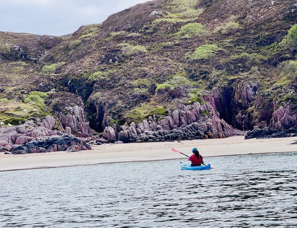paddler with red lifejacket and blue boat approached pink granite and sand beach while sea kayaking in Scotland