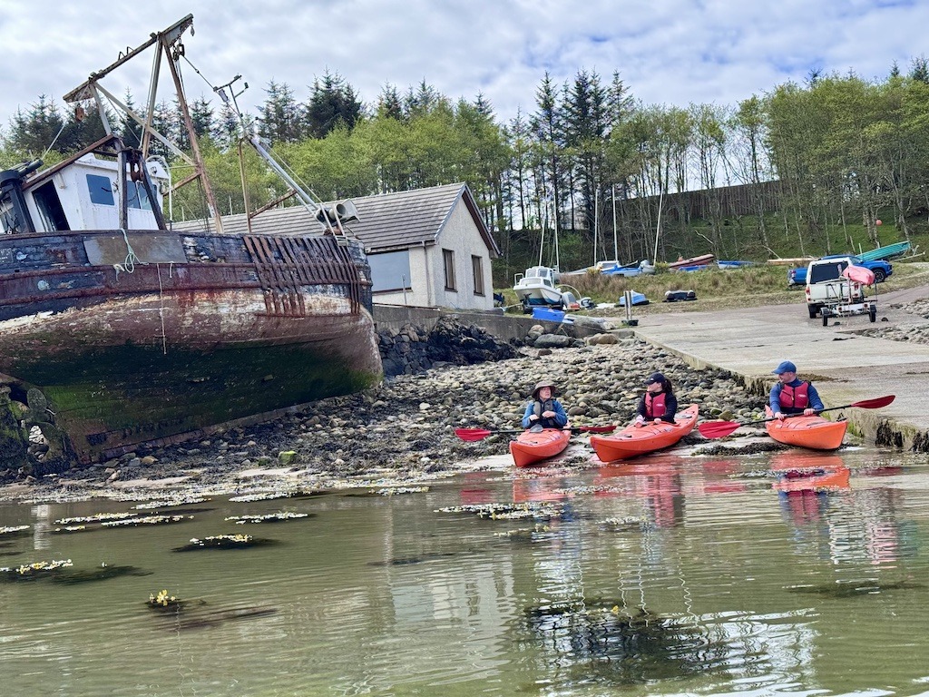 3 people in orange boats next to old ship tipped over a bit-- sea kayaking in Scotland