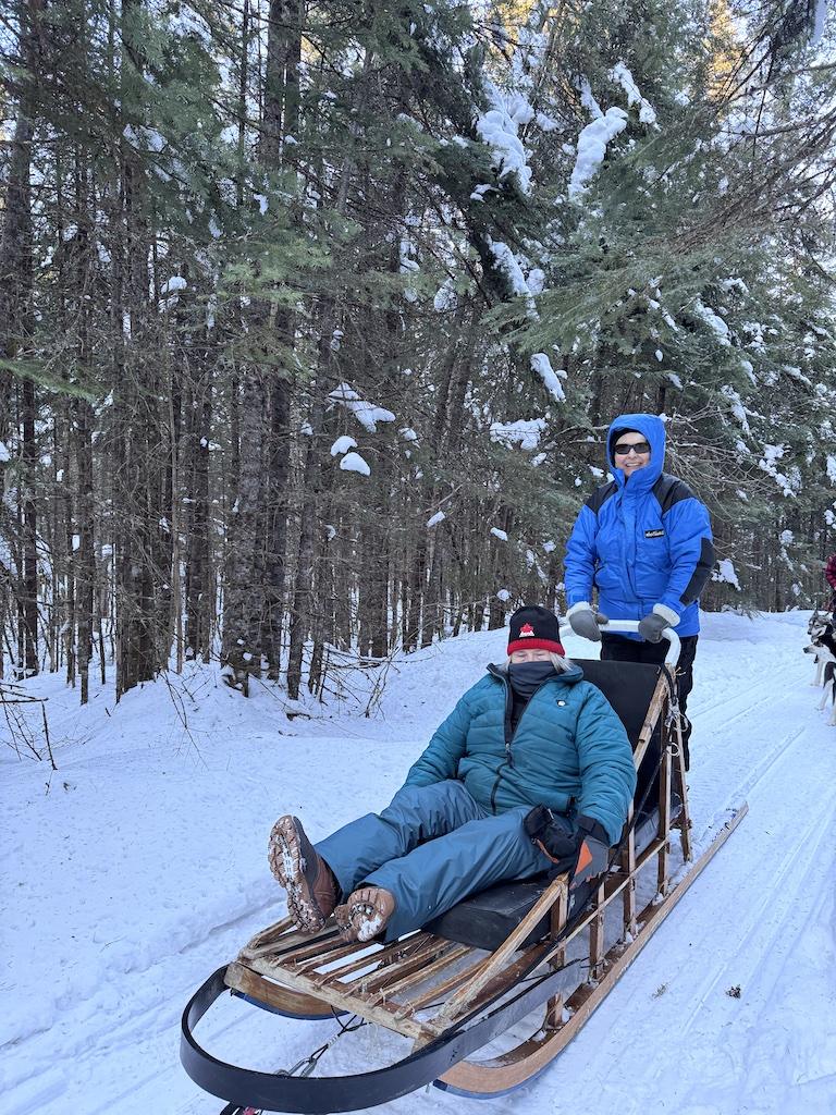 Person with blue coat driving sled while person with green coat rides in the sled while Dogsledding in Quebec
