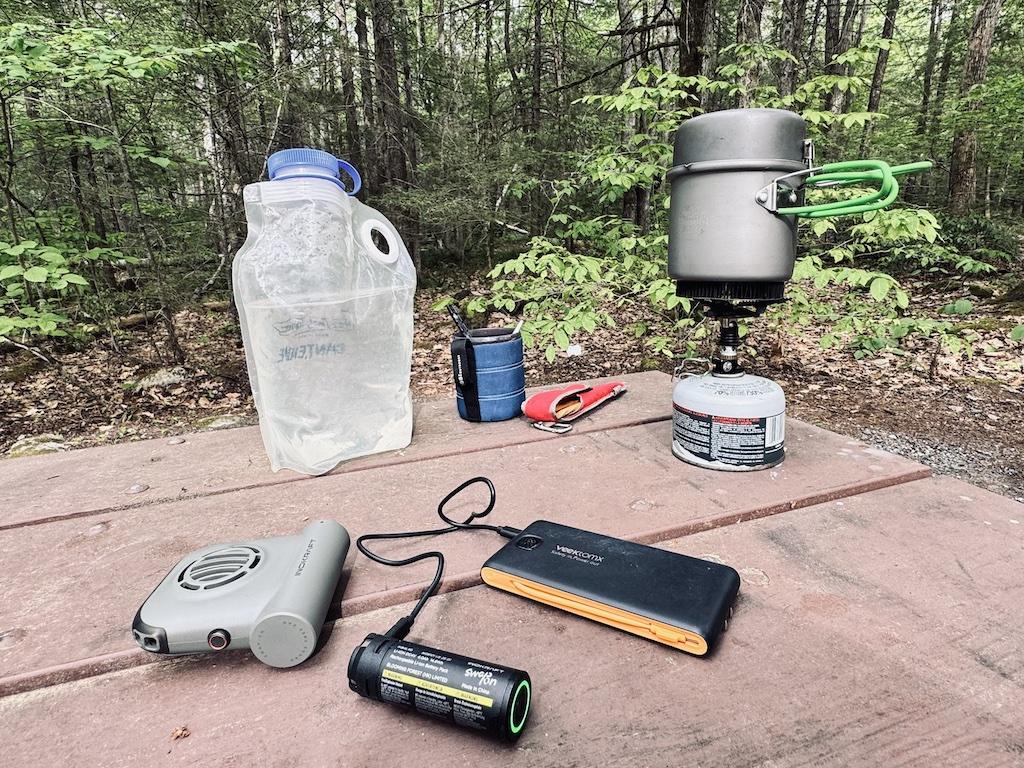trail fan, battery, power bank, camping stove, water bladder, cup and utensils on picnic table