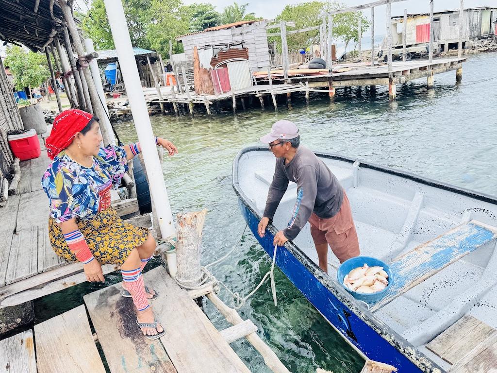 man with fish in a boat while a woman in colorful dress sits on dock