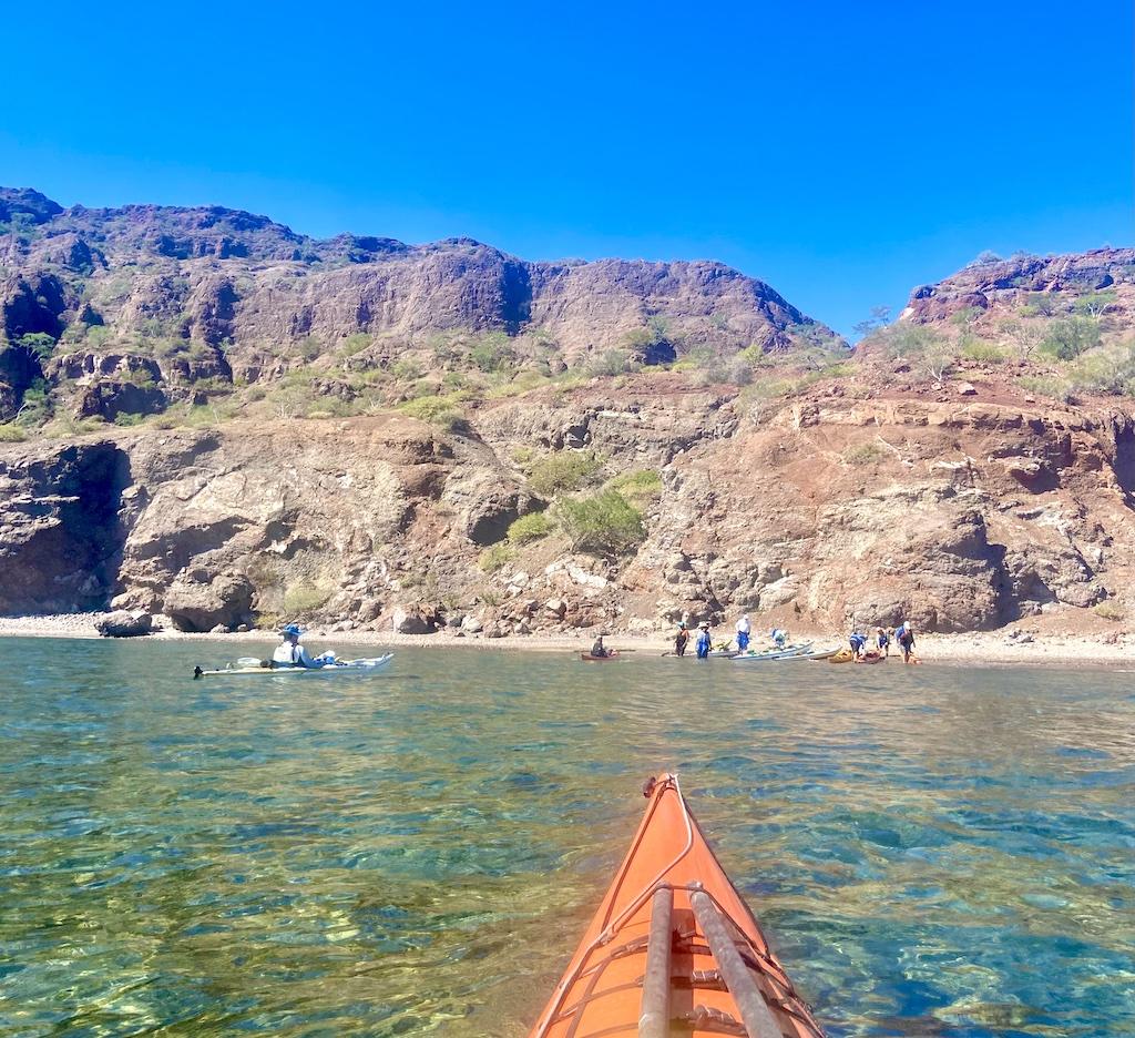 orange kayak approaching a beach with people on an International sea kayaking trip